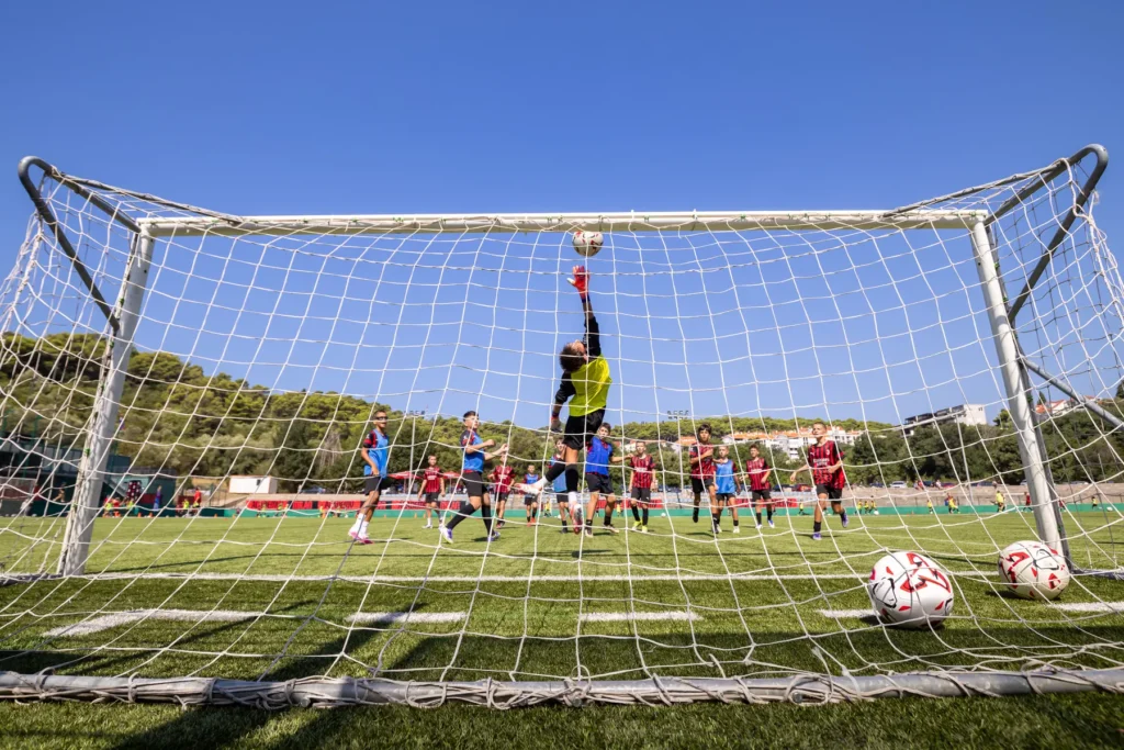 Young goalkeeper jumping to reach the ball at the goal, seen from behind the net on a sunny day