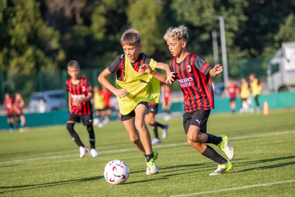 Two boys in a yellow bib and an AC Milan kit chase the ball during a camp training game