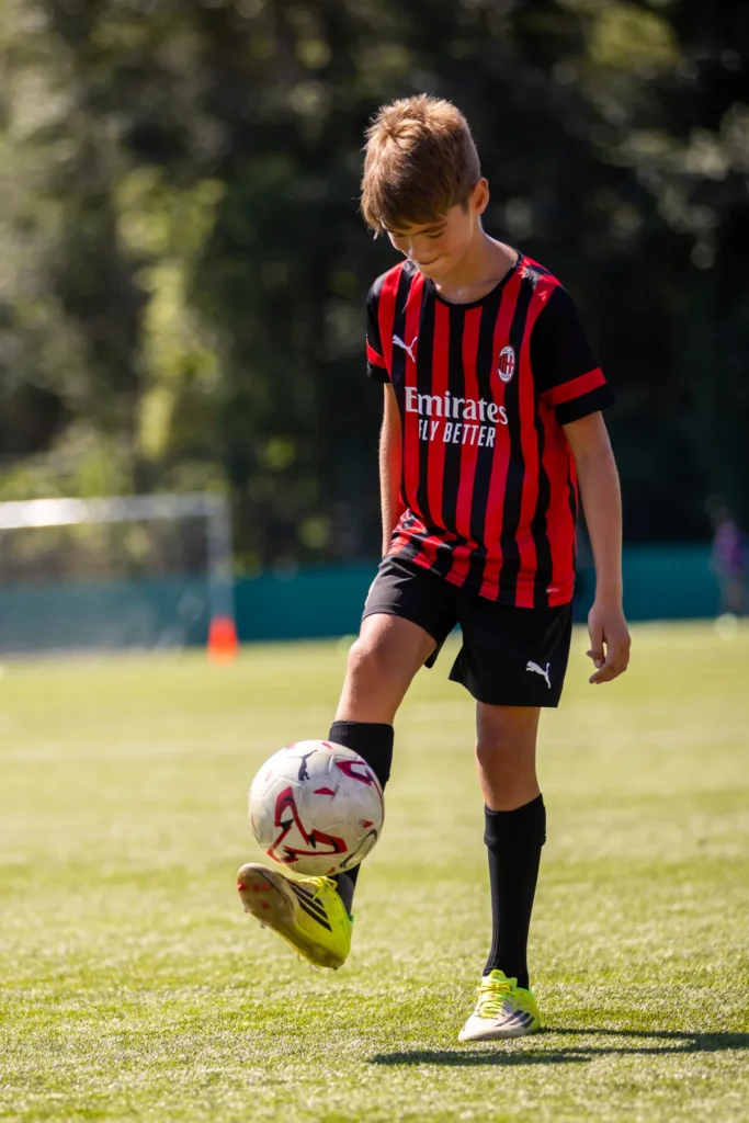 Boy in a red and black AC Milan kit juggling a football on the pitch