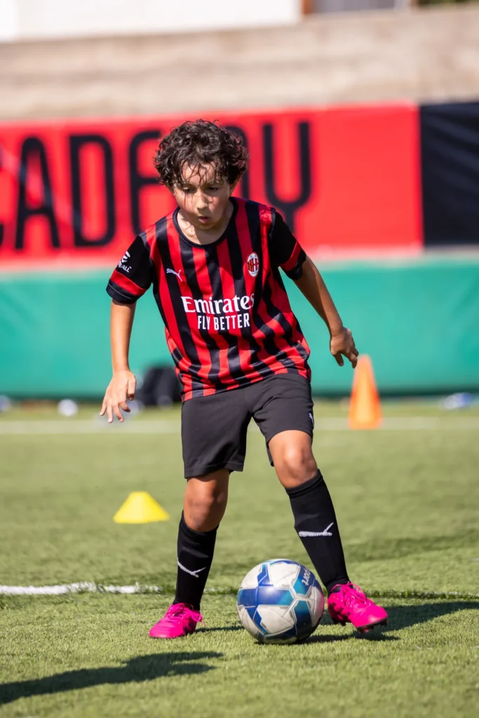 Young player with curly hair dribbling the ball past a yellow cone