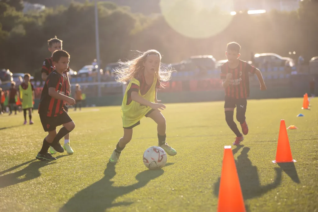 Girl in a yellow bib dribbling past orange cones in warm evening sunlight