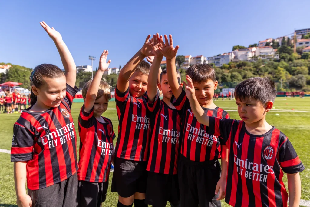 Children in AC Milan kits raising their hands and waving, with a hillside in the background