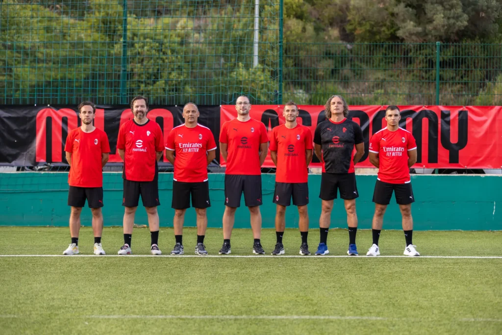 Seven AC Milan Academy coaches in red and black training kit lined up in front of the Milan Academy banner