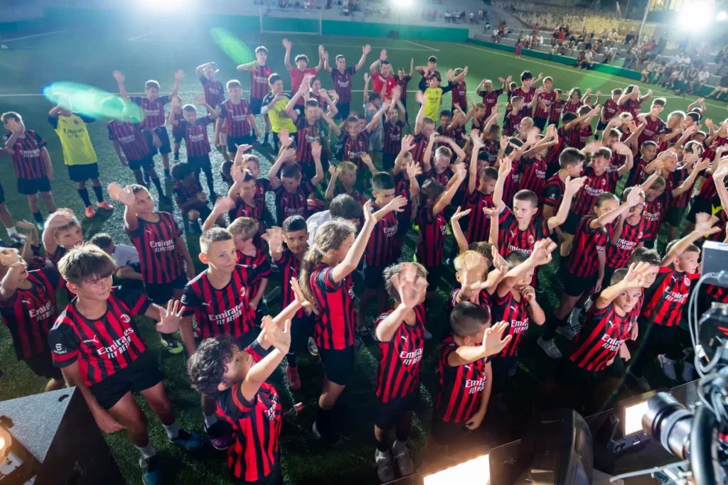 Crowd of children in AC Milan kits raising their arms on the pitch at night under floodlights