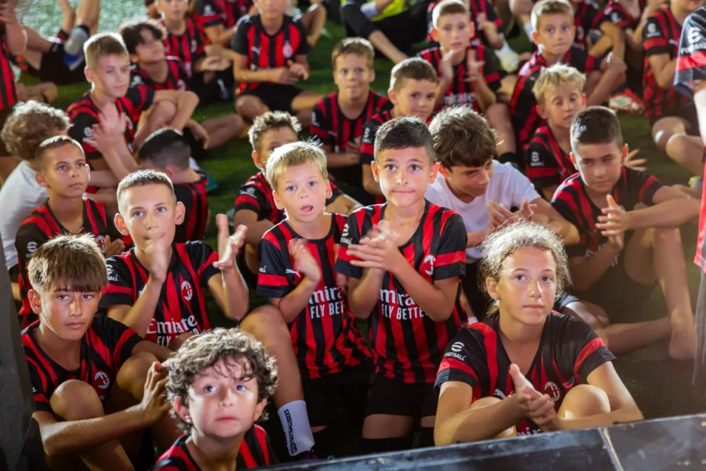 Children in AC Milan kits sitting on the grass, clapping and looking up