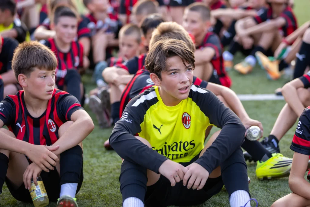 Two boys sitting on the grass, one in a yellow goalkeeper top, watching the training session