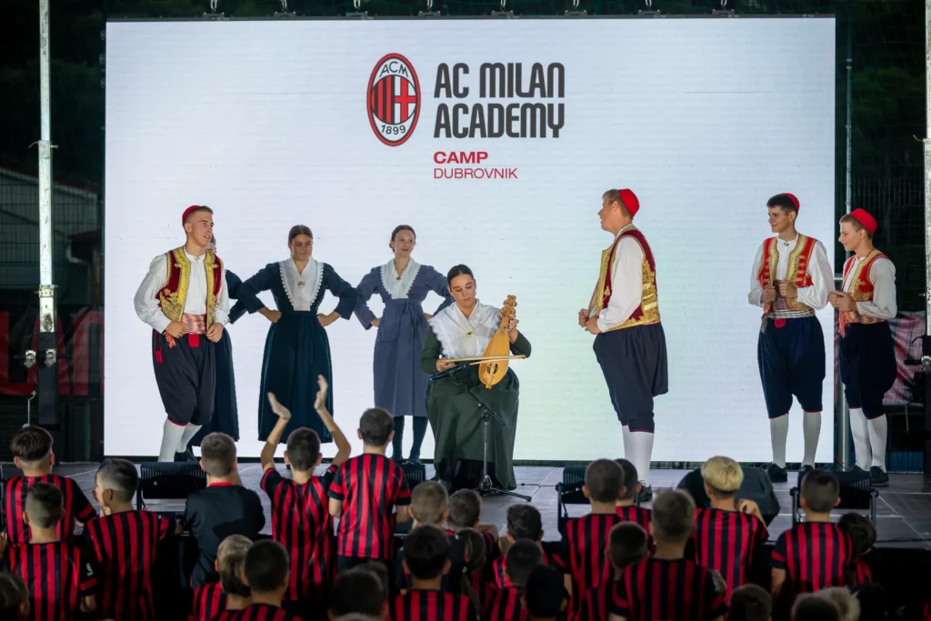 Folklore dancers and a musician performing on stage in front of the AC Milan Academy Camp Dubrovnik screen while children watch